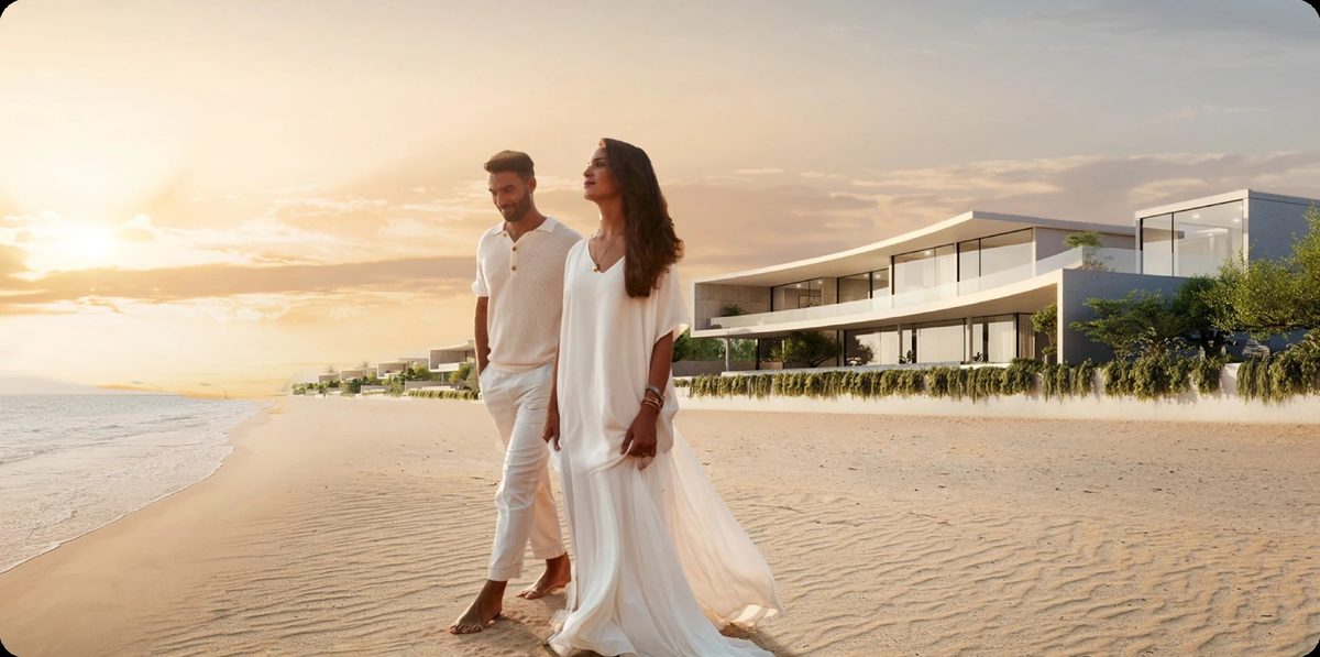 A couple walking along the shoreline at sunset, a row of low-rise beachfront villas behind them